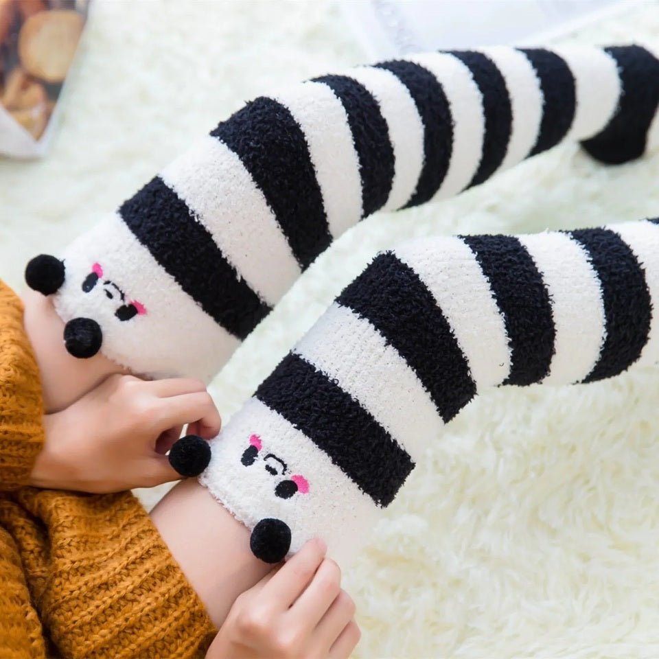 des chaussettes hautes en matière pilou toutes douces rayées noires et blanches. Sur le haut des chaussettes, un visage mignon de panda avec ses joues roses et deux pompons noirs en guise d'oreilles