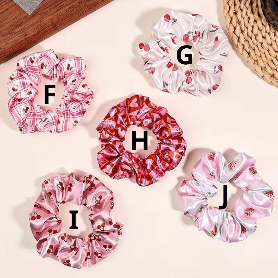 Une collection de chouchous satinés de couleur rose et blanc à motif de cerises. Leur texture brillante est mise en valeur dans un intérieur lumineux aux tons neutres avec un arrière-plan clair.