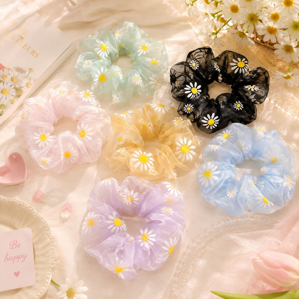 Collection de chouchous en organza à marguerites blanches, déclinés en rose, vert, jaune, bleu, violet et noir, présentés dans une élégante mise en scène fleurie. Accessoires pour cheveux légers et transparents au style romantique, parfaits pour apporter une touche florale et colorée à toutes les coiffures.
