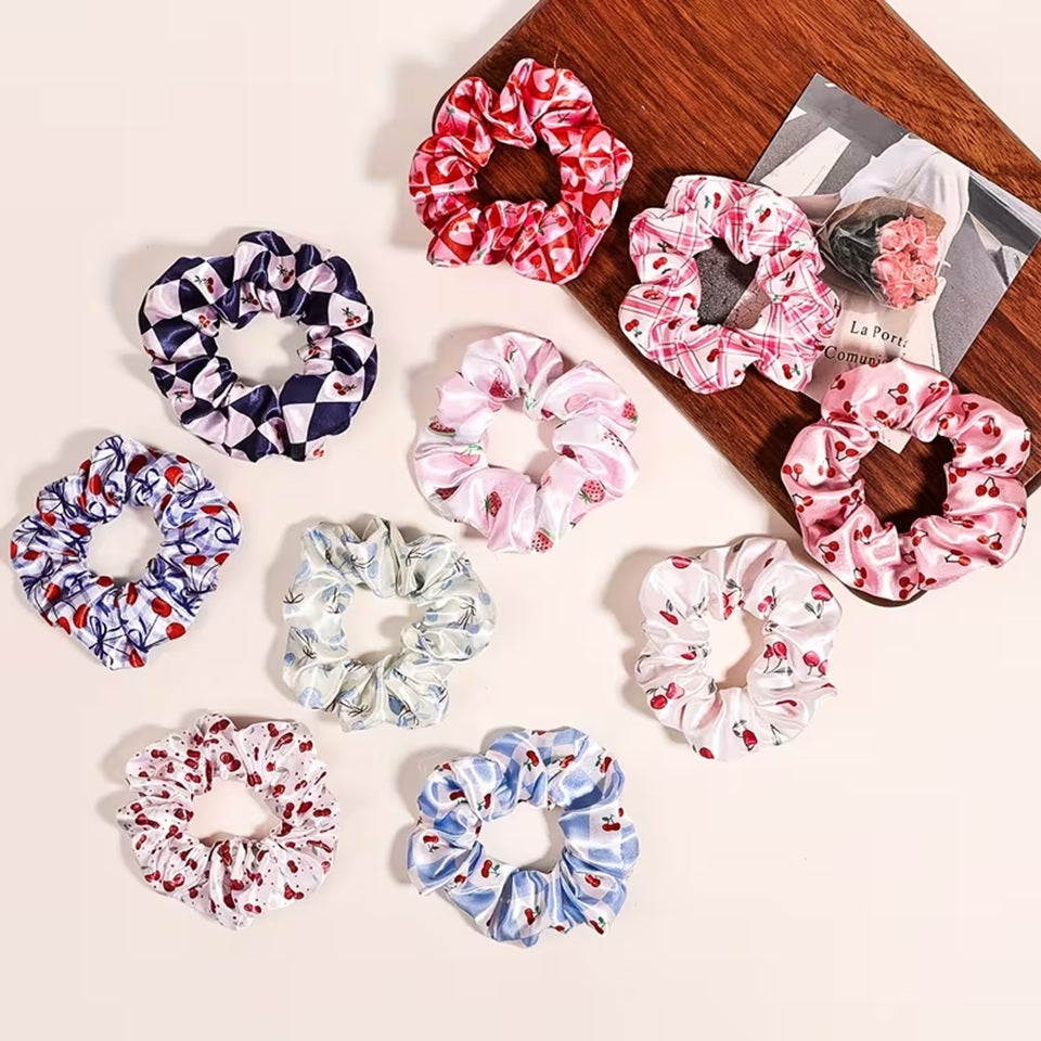 Une collection de chouchous satinés de couleur bleu, rose et blanc à motif de cerises. Leur texture brillante est mise en valeur dans un intérieur lumineux aux tons neutres avec un arrière-plan clair.