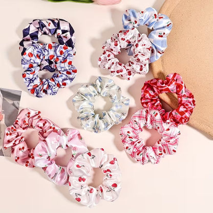 Une collection de chouchous satinés de couleur bleu, rose et blanc à motif de cerises. Leur texture brillante est mise en valeur dans un intérieur lumineux aux tons neutres avec un arrière-plan clair et doux.