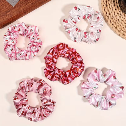 Une collection de chouchous satinés de couleur rose et blanc à motif de cerises. Leur texture brillante est mise en valeur dans un intérieur lumineux aux tons neutres avec un arrière-plan clair.