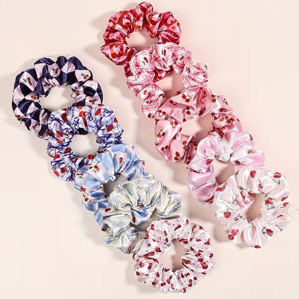 Une collection de chouchous satinés de couleur bleu, rose et blanc à motif de cerises. Leur texture brillante est mise en valeur dans un intérieur lumineux aux tons neutres avec un arrière-plan clair.