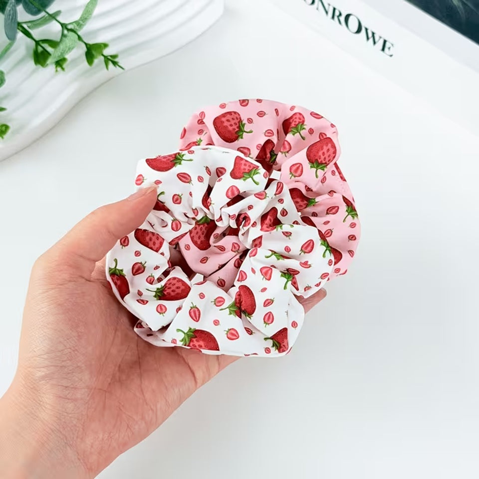 Deux chouchous blanc et rose au motif de fraises rouges gourmandes et fraiches tenus dans une main de femme. Le décor est une table blanche avec des feuilles vertes.