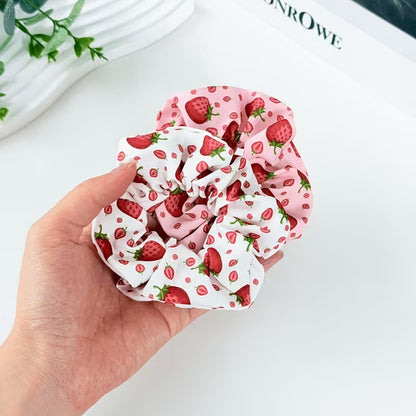Deux chouchous blanc et rose au motif de fraises rouges gourmandes et fraiches tenus dans une main de femme. Le décor est une table blanche avec des feuilles vertes.