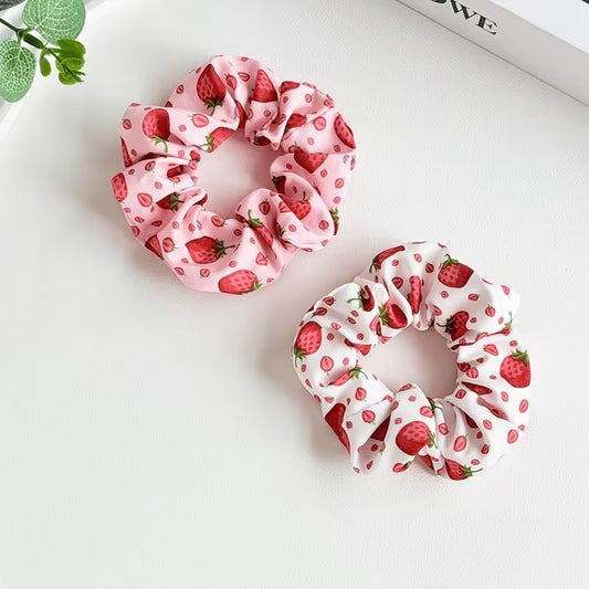 Deux chouchous blanc et rose au motif de fraises rouges gourmandes et fraiches posés sur une table blanche.