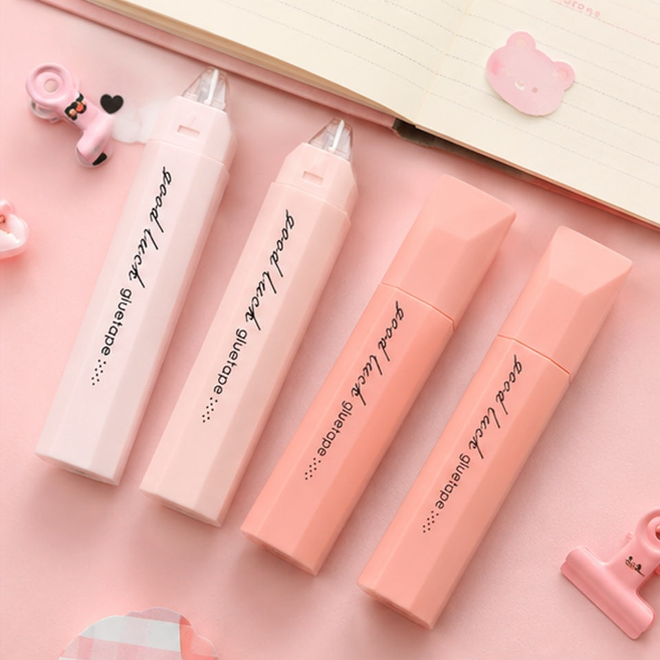 de la colle sous forme de ruban dans leur joli étui rose. Ils sont posés sur un bureau mignon.