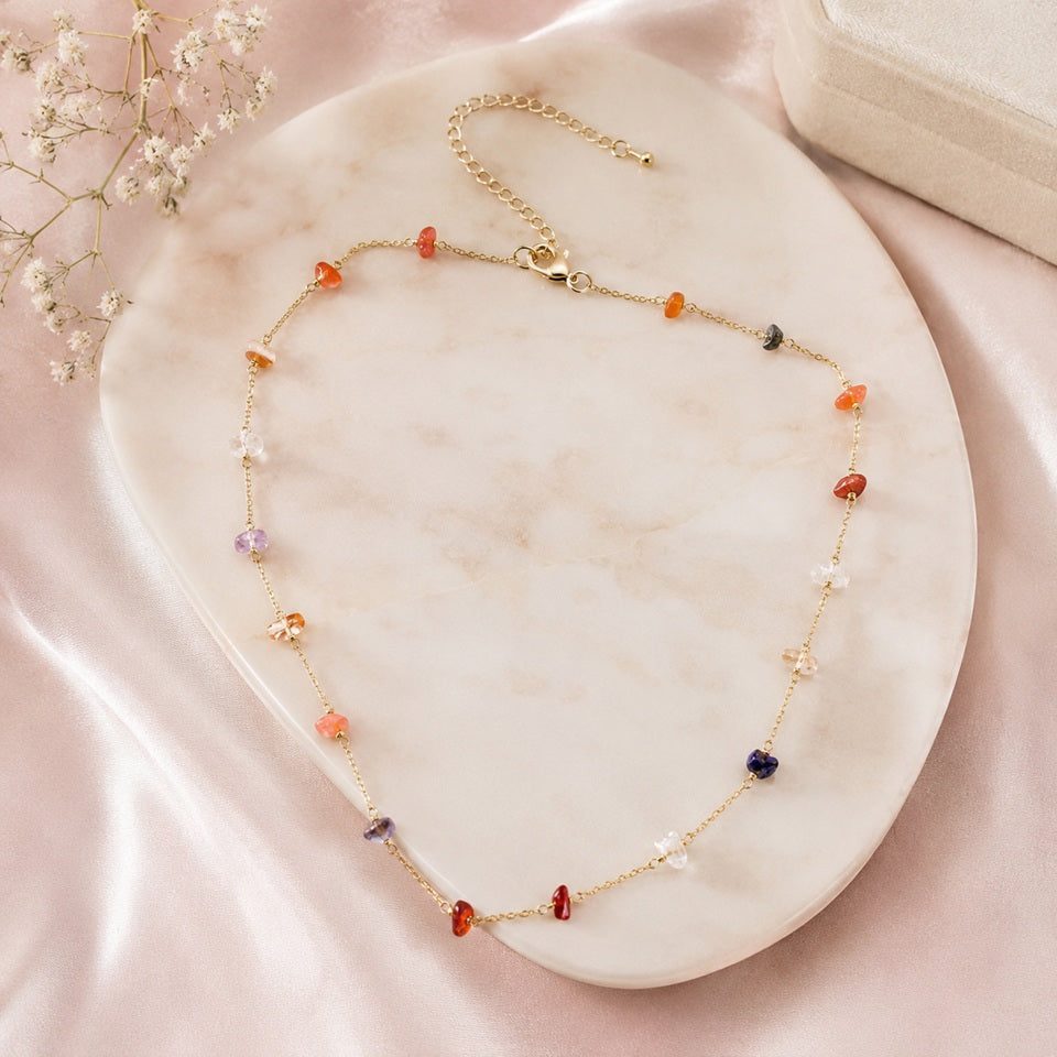 un collier doré fin orné de petites pierres colorées, présenté sur un plateau en marbre clair entouré de satin rose et de fleurs blanches.