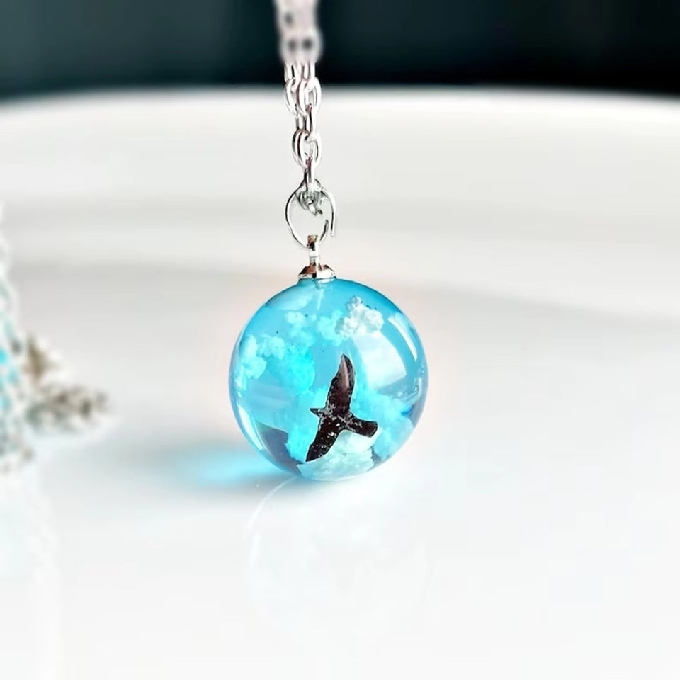 un joli collier argenté d'une sphère transparente et bleue renfermant un oiseau noir volant parmi des nuages comme si un bout de ciel y avait été capturé.
