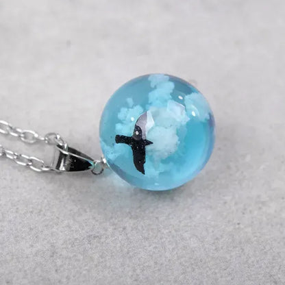un joli collier argenté d'une sphère transparente et bleue renfermant un oiseau noir volant parmi des nuages comme si un bout de ciel y avait été capturé.