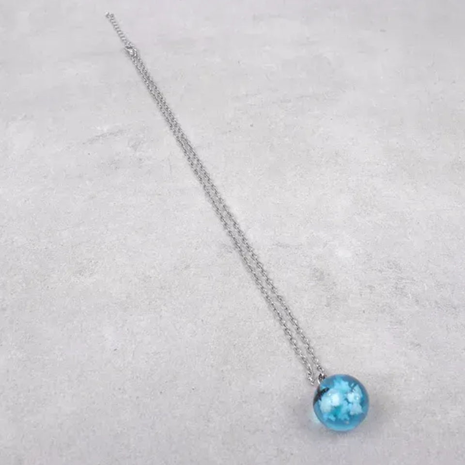 un joli collier argenté d'une sphère transparente et bleue renfermant un oiseau noir volant parmi des nuages comme si un bout de ciel y avait été capturé.