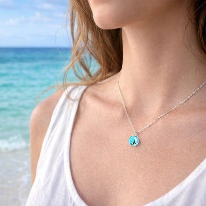 un joli collier argenté d'une sphère transparente et bleue renfermant un oiseau noir volant parmi des nuages comme si un bout de ciel y avait été capturé.