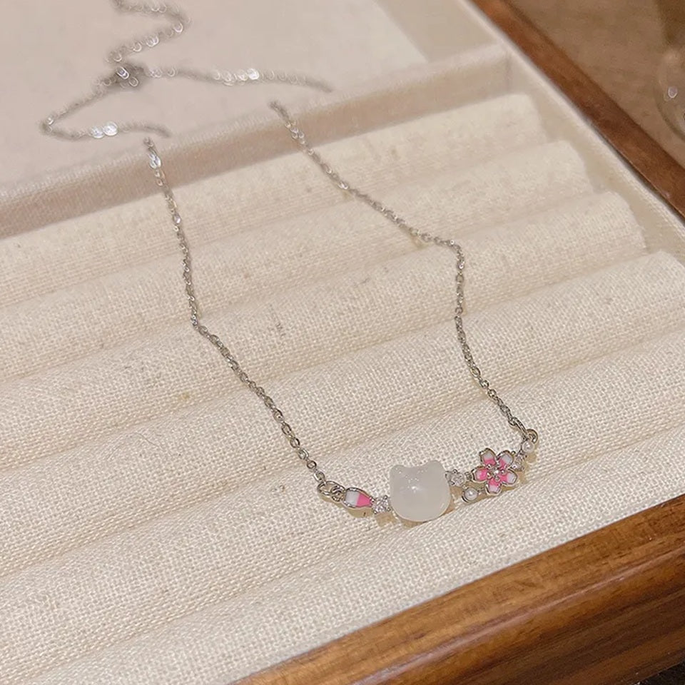 un superbe collier en acier inoxydable argenté composé d'une pierre blanche sculptée en forme de tête de chat, d'une fleur de sakura et d'un pétale rose et de petites perles blanches. L'ensemble est poétique, raffiné et plein de poésie.