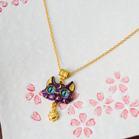 un collier doré dont le pendentif est un chat violet aux grands yeux bleus expressifs. Il a des décorations dorées comme une breloque en forme de soleil.