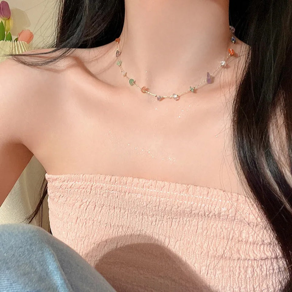 un collier doré fin orné de petites pierres colorées, porté par une femme. Ambiance douce et chaleureuse. Style mignon, moderne et plein de poésie