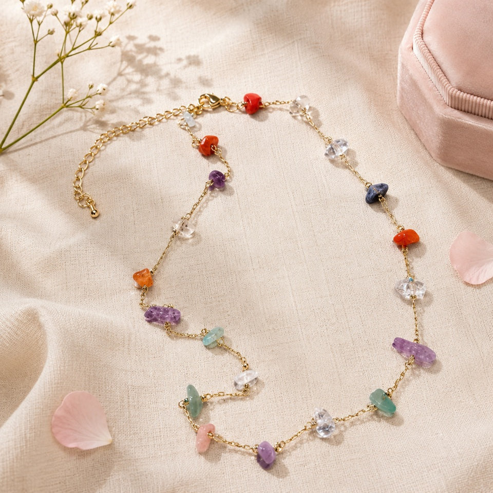 un collier doré orné de pierres naturelles colorées, présenté sur un tissu beige avec des pétales roses, des fleurs blanches et un écrin en velours.