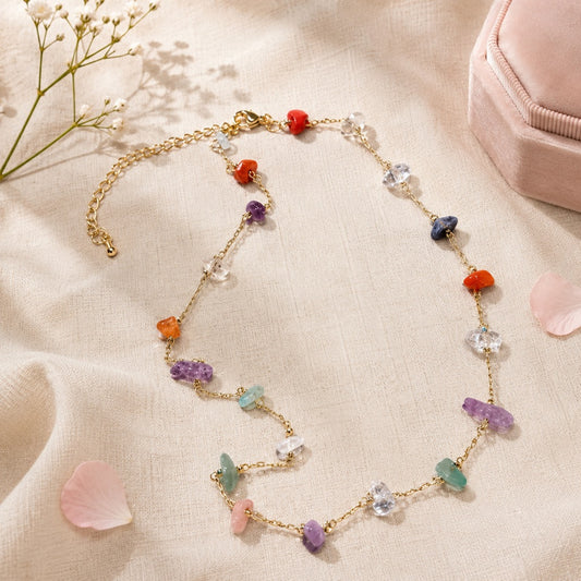 un collier doré orné de pierres naturelles colorées, présenté sur un tissu beige avec des pétales roses, des fleurs blanches et un écrin en velours.