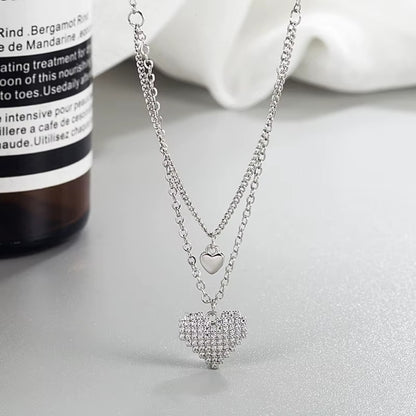 un collier composé de deux chaines reliées. Deux pendentifs en forme de coeur sont les chaines, dont un tout en strass brillants.