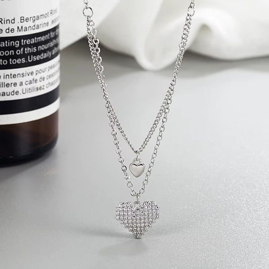 un collier composé de deux chaines reliées. Deux pendentifs en forme de coeur sont les chaines, dont un tout en strass brillants.