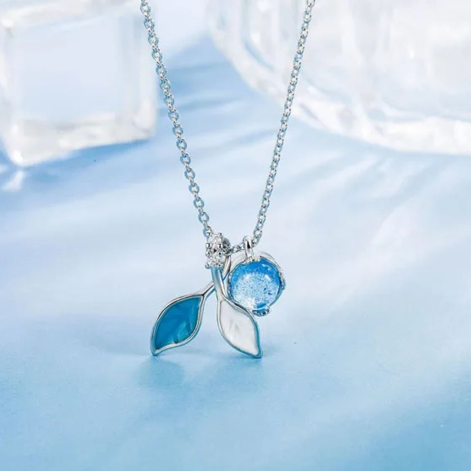 un collier argenté dont le pendentif représente de délicates feuilles bleues et d'une perle aux reflets bleutées. L'ensemble est poétique.