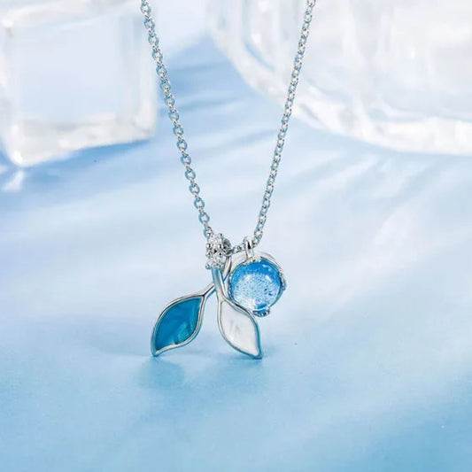 un collier argenté dont le pendentif représente de délicates feuilles bleues et d'une perle aux reflets bleutées. L'ensemble est poétique.