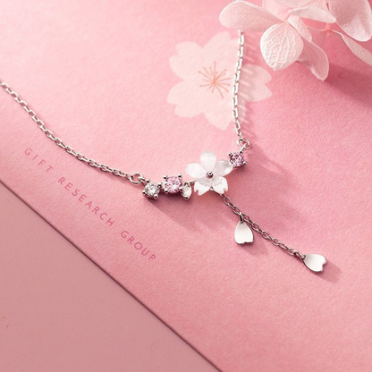 un collier argenté au design d'une délicate fleur de sakura blanche d'où pend une chainette ornée de pétales de saura. Autour de la fleur, il y a des petits strass blancs et roses qui ajoutent de la brillance.