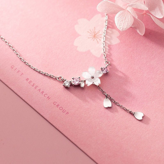 un collier argenté au design d'une délicate fleur de sakura blanche d'où pend une chainette ornée de pétales de saura. Autour de la fleur, il y a des petits strass blancs et roses qui ajoutent de la brillance.