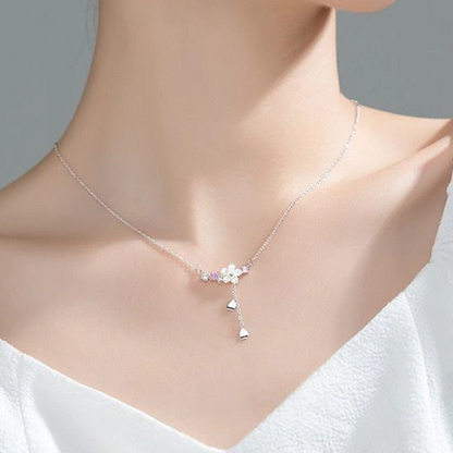 un collier argenté au design d'une délicate fleur de sakura blanche d'où pend une chainette ornée de pétales de saura. Autour de la fleur, il y a des petits strass blancs et roses qui ajoutent de la brillance.