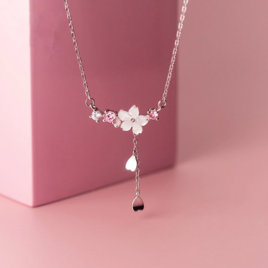 un collier argenté au design d'une délicate fleur de sakura blanche d'où pend une chainette ornée de pétales de saura. Autour de la fleur, il y a des petits strass blancs et roses qui ajoutent de la brillance.