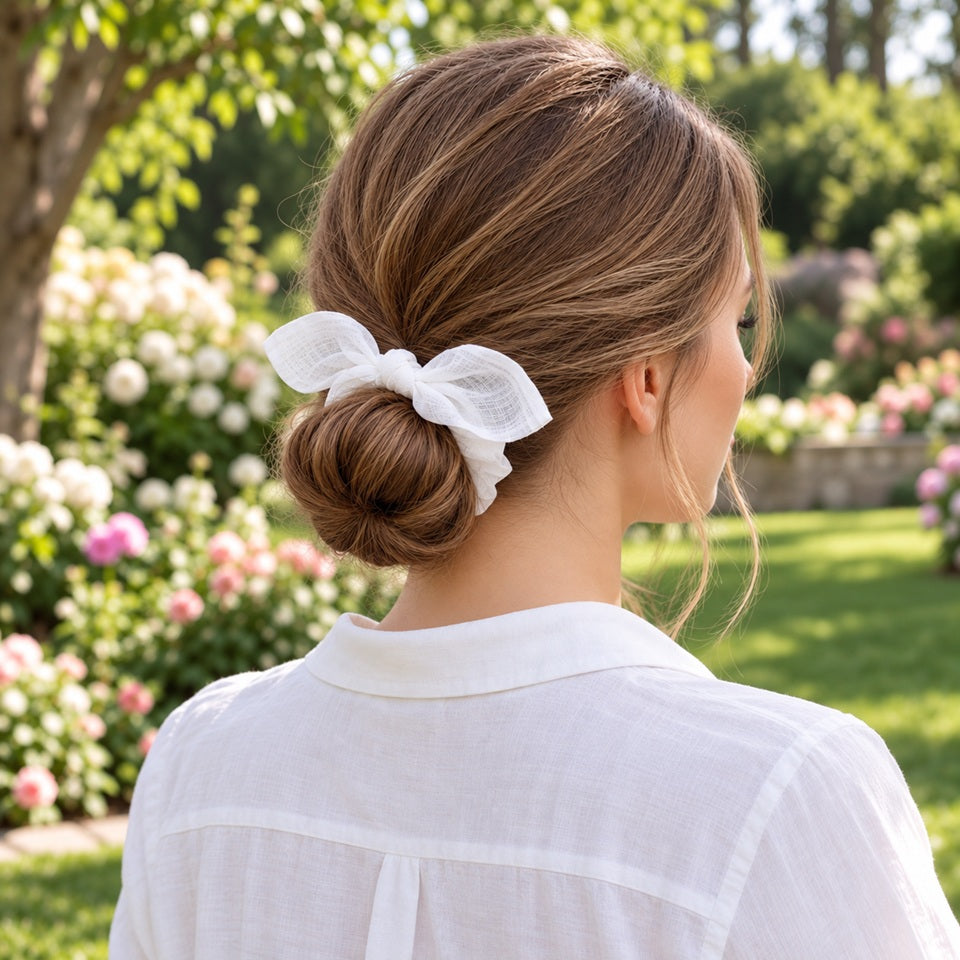 Chouchou blanc avec grand nœud porté sur un chignon par une femme en chemise blanche, dans un jardin fleuri élégant et romantique.