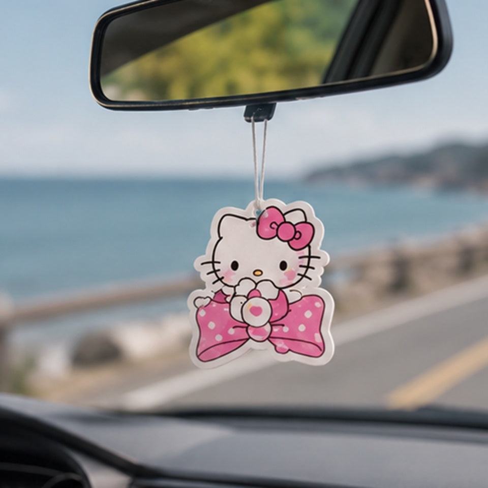 un désodorisant en forme de hello kitty avec son noeud rose emblématique suspendu à un rétroviseur intérieur d'une voiture, qui roule sur une route ensoleillée.