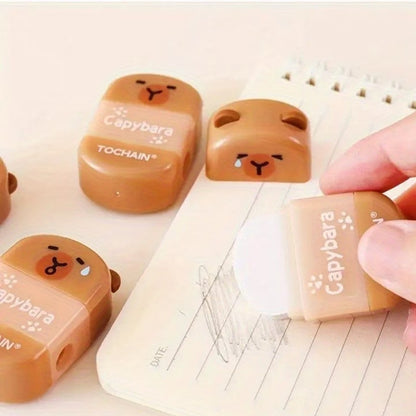la gomme capybara est tenue par une femme en train d'effacer du crayon sur un carnet. D'autres gommes et taille-crayons capybara sont posés autour.