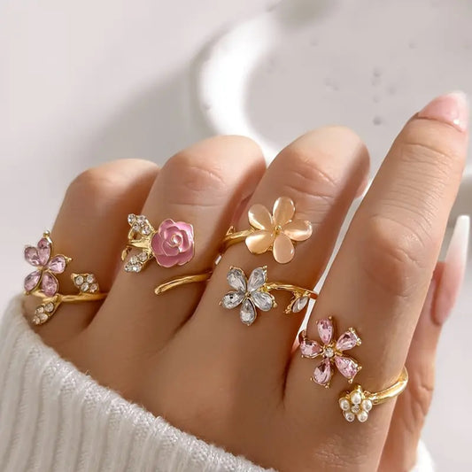 un ensemble de 4 bagues en alliage doré au design de fleurs roses élégantes et de strass brillants roses et argentés avec des perles. L'ensemble est romantique et féminin