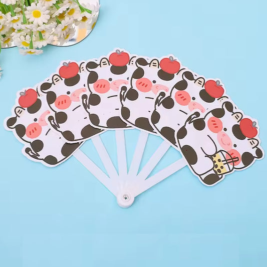 un éventail dont les lames représentent de mignonnes vaches noires et blanches qui boivent un bubble tea et qui ont une pomme rouge sur la tête. L'ensemble est adorale, c'est un accessoire utile et pratique pour les chaudes journées.