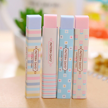 Quatre jolies gommes rectangulaires rose et bleu au motif de lignes colorées et de fleurs blanches vues de face, posées sur un bureau.