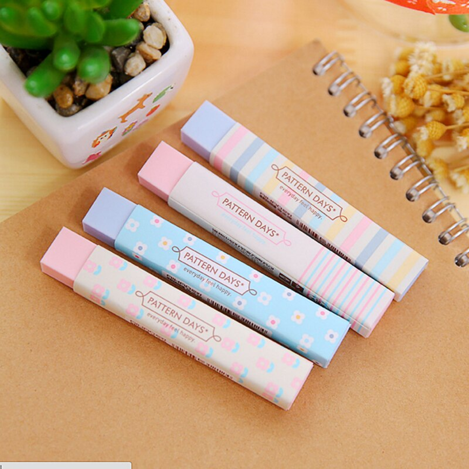 Quatre jolies gommes rectangulaires rose et bleu au motif de lignes colorées et de fleurs blanches posées sur un carnet marron dans un décor naturel et calme.