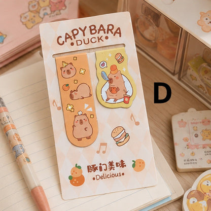 Deux marque-pages magnétiques capybara kawaii orange et jaune, présentés sur un carnet dans un joli décor de papeterie pastel.