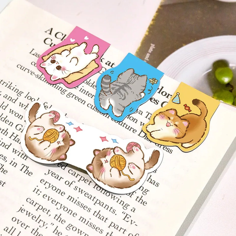 Des marque-pages aimantés en forme de chats gris, blanc, roux et brun accrochés à une page d'un livre. Ils sont adorables dans leurs poses rigolotes et pleines de vie.