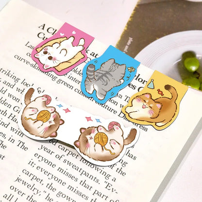 Des marque-pages aimantés en forme de chats gris, blanc, roux et brun accrochés à une page d'un livre. Ils sont adorables dans leurs poses rigolotes et pleines de vie.