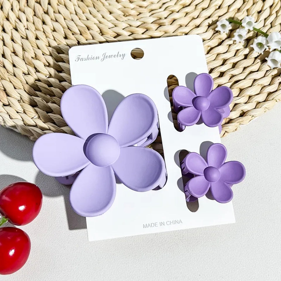 un lot de 2 petites pinces et 1 grande pince à cheveux mauve en forme de fleur joliment présentées sur un support blanc posé sur un tapis rond en rotin. Ambiance estival, lumineuse.
