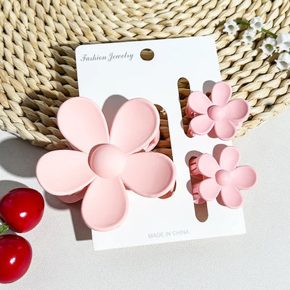 un lot de 2 petites pinces et 1 grande pince à cheveux rose en forme de fleur joliment présentées sur un support blanc posé sur un tapis rond en rotin. Ambiance estival, lumineuse.