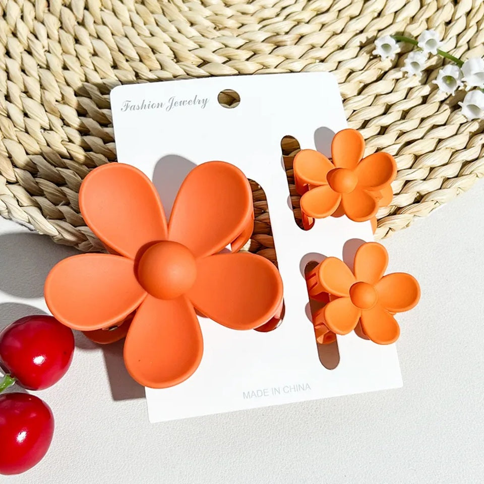 un lot de 2 petites pinces et 1 grande pince à cheveux orange en forme de fleur joliment présentées sur un support blanc posé sur un tapis rond en rotin. Ambiance estival, lumineuse.