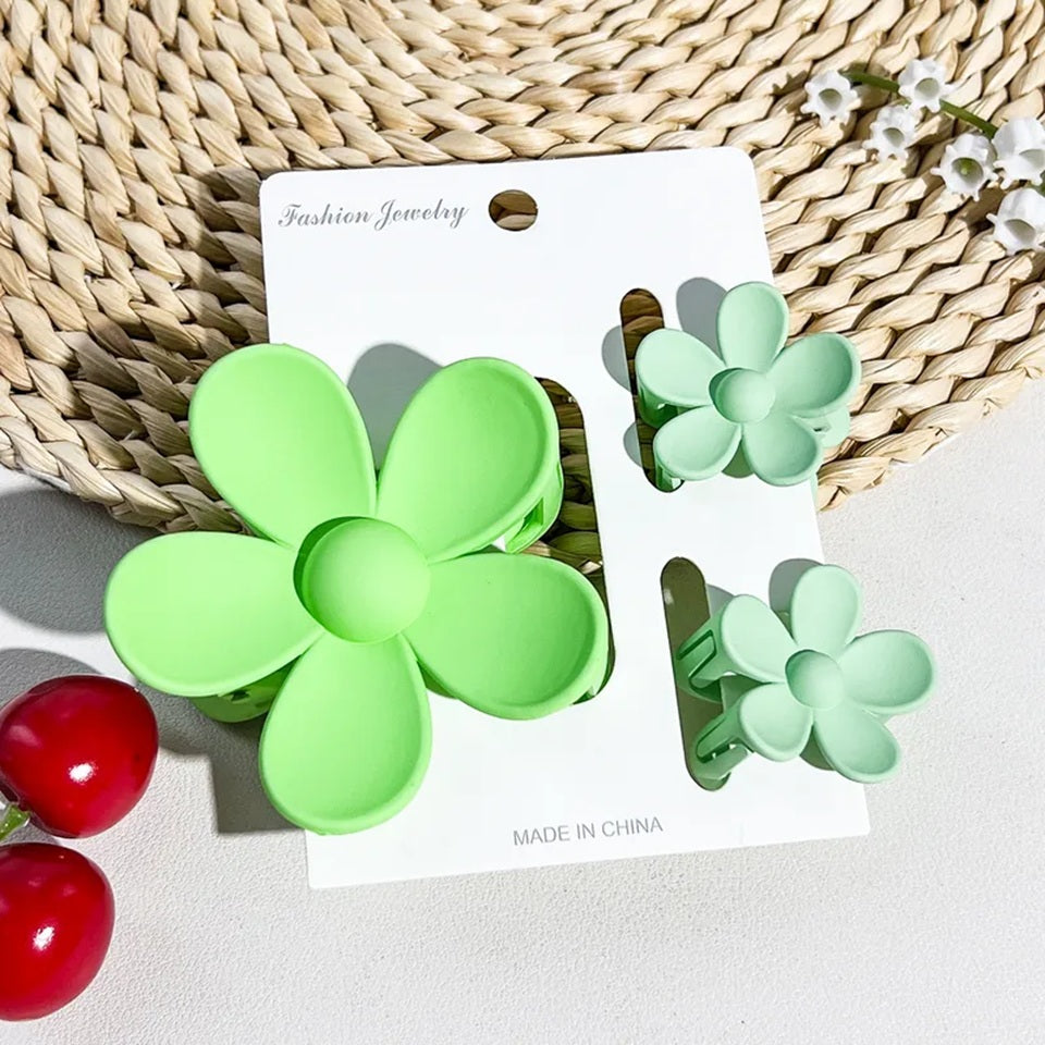 un lot de 2 petites pinces et 1 grande pince à cheveux vert en forme de fleur joliment présentées sur un support blanc posé sur un tapis rond en rotin. Ambiance estival, lumineuse.