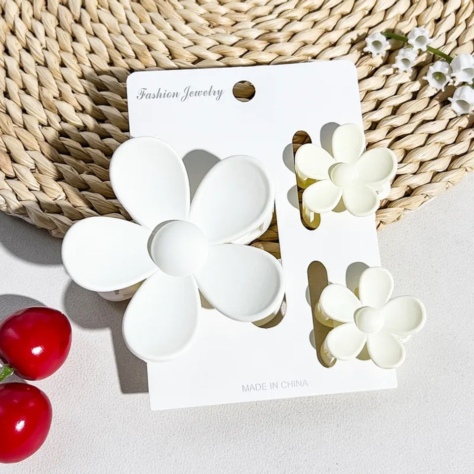 un lot de 2 petites pinces et 1 grande pince à cheveux blanc en forme de fleur joliment présentées sur un support blanc posé sur un tapis rond en rotin. Ambiance estival, lumineuse.