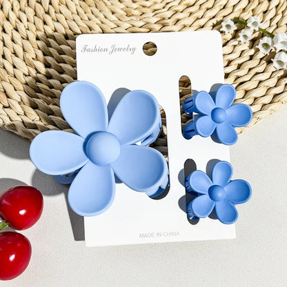 un lot de 2 petites pinces et 1 grande pince à cheveux bleu en forme de fleur joliment présentées sur un support blanc posé sur un tapis rond en rotin. Ambiance estival, lumineuse.