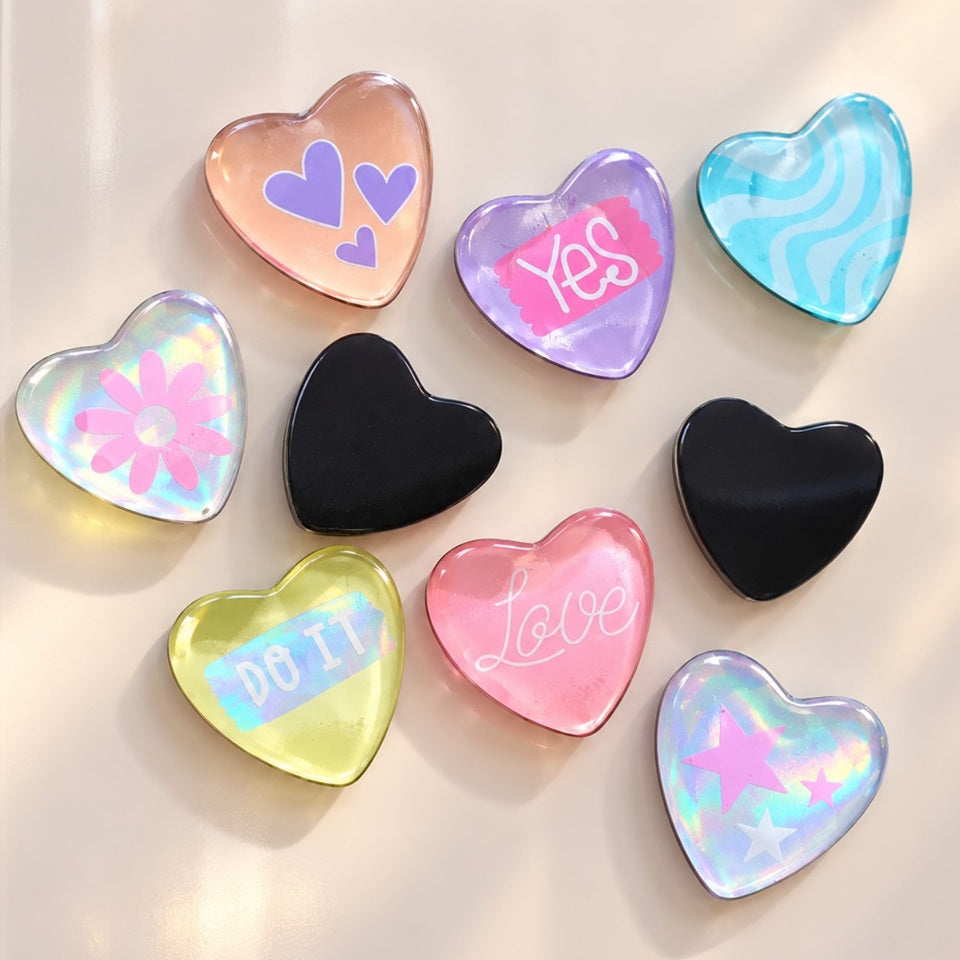 Lot de neuf magnets kawaii en forme de cœur, aux couleurs pastel et aux finitions brillantes, présentés sur un réfrigérateur blanc. Ces adorables aimants décoratifs présentent des motifs variés : inscriptions « Love », « Yes » et « DO IT », petits cœurs violets, fleur rose, étoiles holographiques, vagues turquoise et deux cœurs noirs unis. Leurs effets transparents, irisés et scintillants apportent une touche de douceur et de fantaisie. 