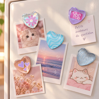 Cinq magnets kawaii en forme de cœur, aux couleurs pastel et aux finitions brillantes, disposés sur un tableau magnétique blanc pour maintenir de jolies photos et petits mots doux. Ces adorables aimants décoratifs présentent différents motifs : fleur rose sur fond holographique, inscription « Yes » sur cœur violet, vagues turquoise, petits cœurs mauves et effet pailleté argenté. Ils maintiennent des photos.