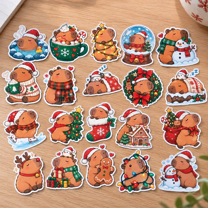 Lot de stickers capybara de Noël kawaii avec bonnets, sapins, cadeaux, bonhommes de neige et décorations festives, disposés sur un bureau en bois clair.