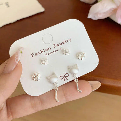 un lot de 3 paires de boucles d'oreilles argentées sur le thème des chats : deux paires de puces en forme de tête avec un strass et de patte de chat. Une paire de boucles d'oreilles en forme de tête de chat d'où pend une adorable queue de chat. L'ensemble est dans un style minimaliste et adorable.
