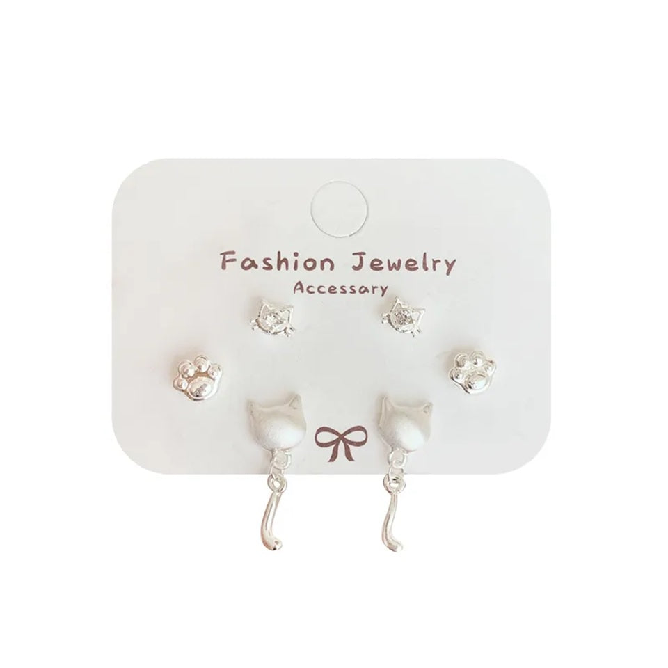 un lot de 3 paires de boucles d'oreilles argentées sur le thème des chats : deux paires de puces en forme de tête avec un strass et de patte de chat. Une paire de boucles d'oreilles en forme de tête de chat d'où pend une adorable queue de chat. L'ensemble est dans un style minimaliste et adorable.